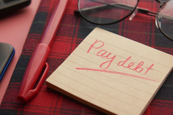 Pay debt reminder note with pen and glasses on plaid surface