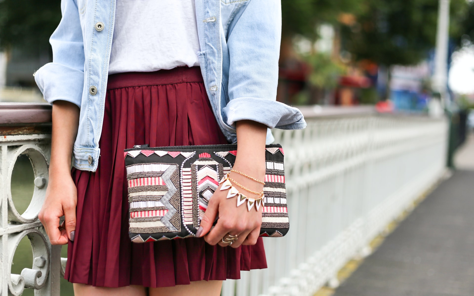 Stylish woman in denim jacket and pleated skirt with a patterned clutch and bracelets.